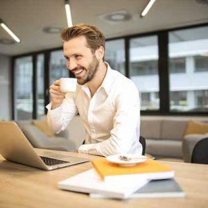 Ein Mann sitz vor seinem Laptop und trinkt mit einem Lächeln sienen Kaffee